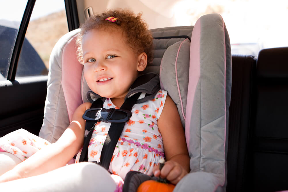 A young child wearing a white dress with colorful floral patterns sits in a grey car seat with pink accents inside a vehicle. 