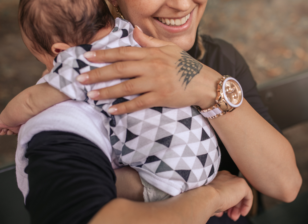 Une personne tient un bébé vêtu d’une tenue géométrique blanche et grise. La personne a un tatouage sur la main, porte une montre-bracelet et sourit à moitié.