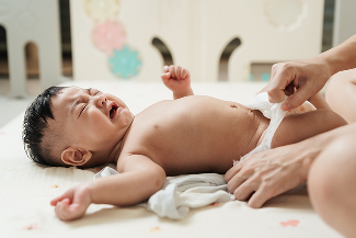 A baby lying down and crying as an adult reaches to undo their diaper.