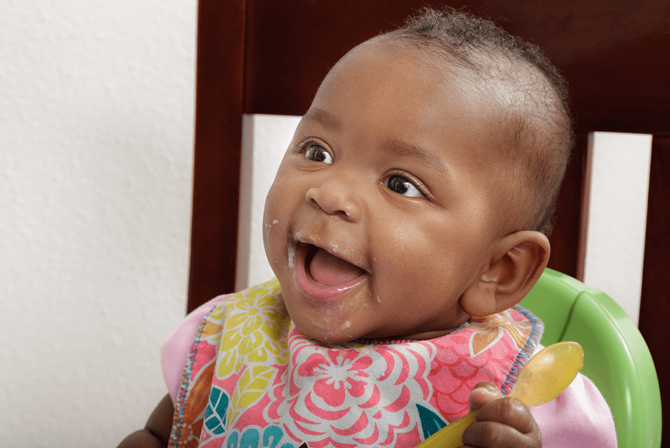 Un bébé assis dans une chaise haute et portant un bavoir coloré tient une cuillère de couleur jaune.