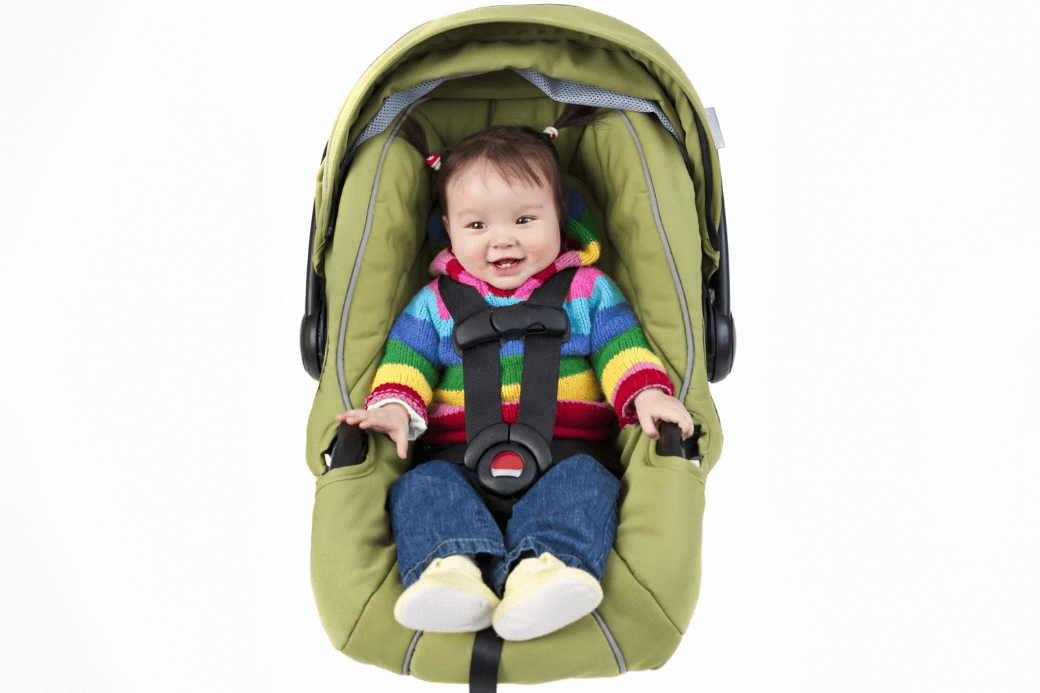 A baby wearing a colorful striped sweater, blue jeans, and yellow shoes is seated in a green car seat with black safety straps.