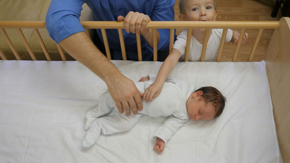 Un bébé en grenouillère dort sur le dos dans un lit de bébé. Un adulte et un tout-petit étirent un bras pour le toucher doucement