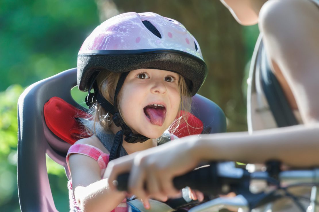 A child in a pink polka-dot helmet sits in a bike seat, with an adult holding the handlebars. The scene is outdoors with greenery.