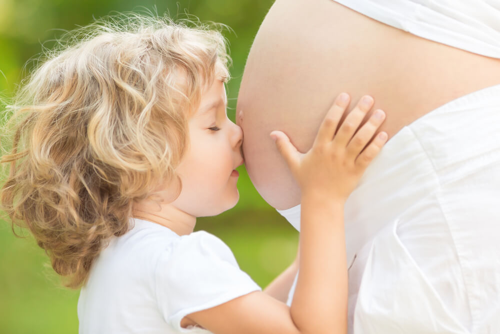 Un jeune enfant aux cheveux blonds touche le ventre d’une personne enceinte. L’arrière-plan est vert et flou, ce qui indique que le décor est extérieur.