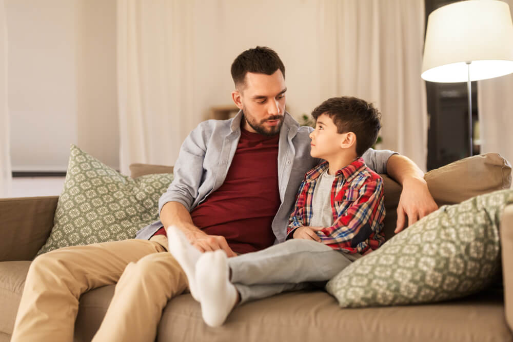 Un adulte et un enfant s’assoient sur le canapé, dans un salon. L’adulte, qui porte une chemise bleu pâle par-dessus un t-shirt rouge et un pantalon beige, a un bras autour de l’enfant, qui porte une chemise à carreaux rouge et bleu et un pantalon gris. L’adulte et l’enfant se regardent. Des oreillers à motifs et une lampe sur pied sont visibles en arrière-plan.