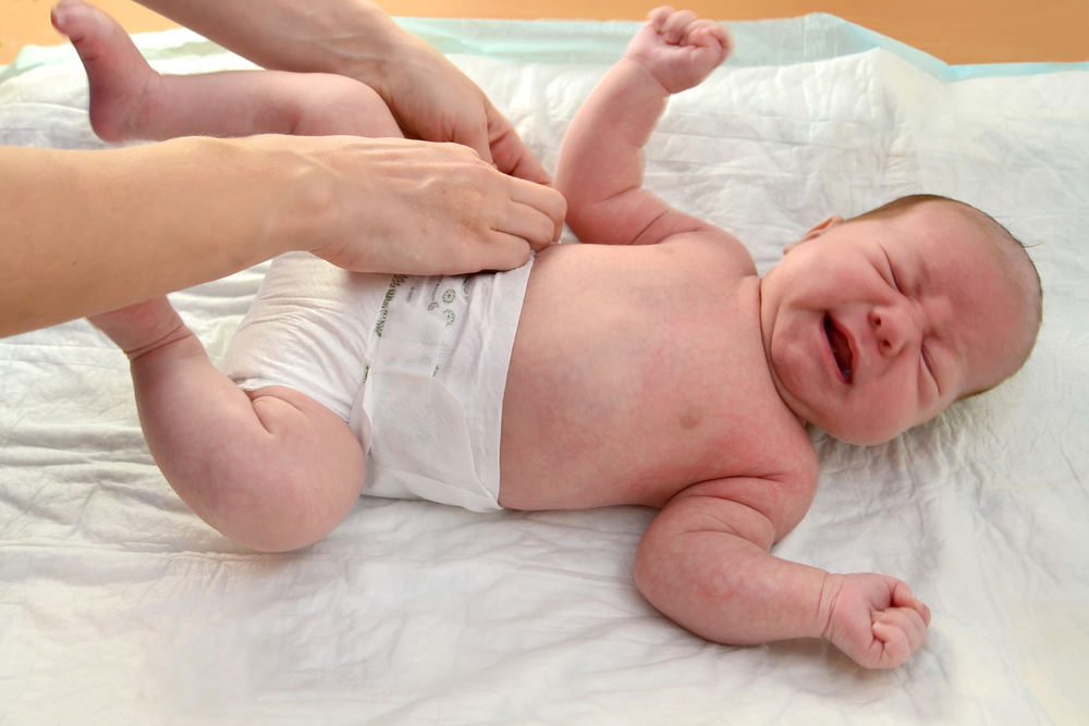 A baby lying down and crying as an adult reaches to undo their diaper.