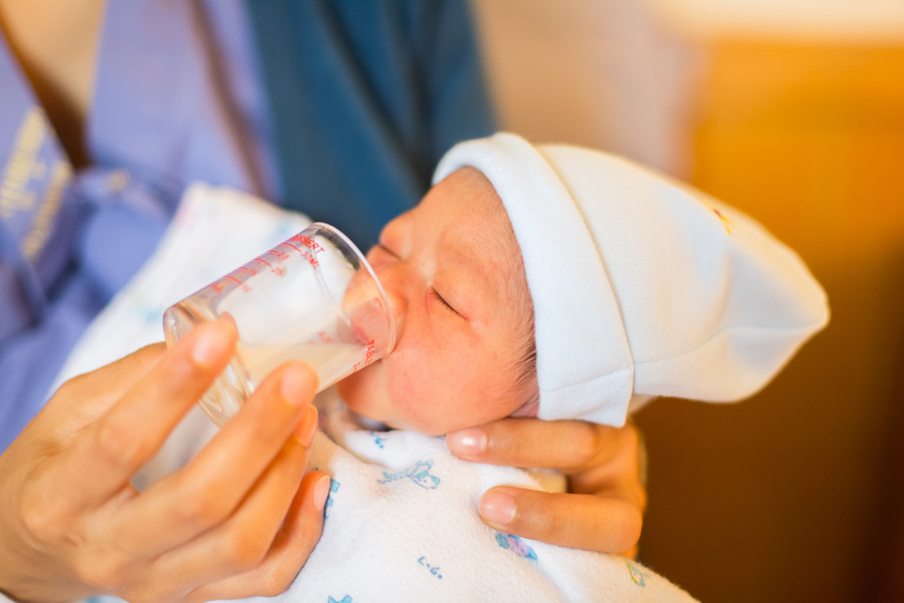 A newborn baby in a white hat and blanket is fed milk from a small measuring cup by an adult
