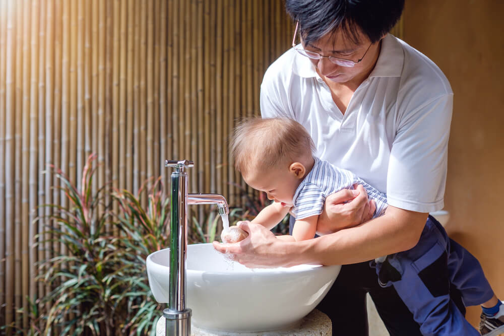 Adulte tenant un bébé au-dessus d’un lavabo blanc, aidant le bébé à se laver les mains sous l’eau courante d’un robinet. L’adulte porte une chemise blanche, et le bébé est vêtu d’une tenue à rayures bleues. En arrière-plan, on voit des bâtons de bambou verticaux et quelques plantes.