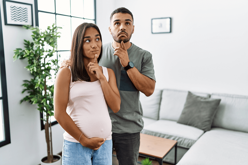 Two people stand in a modern living room, both making a thinking gesture, with an index finger to the chin. One person is visibly pregnant.