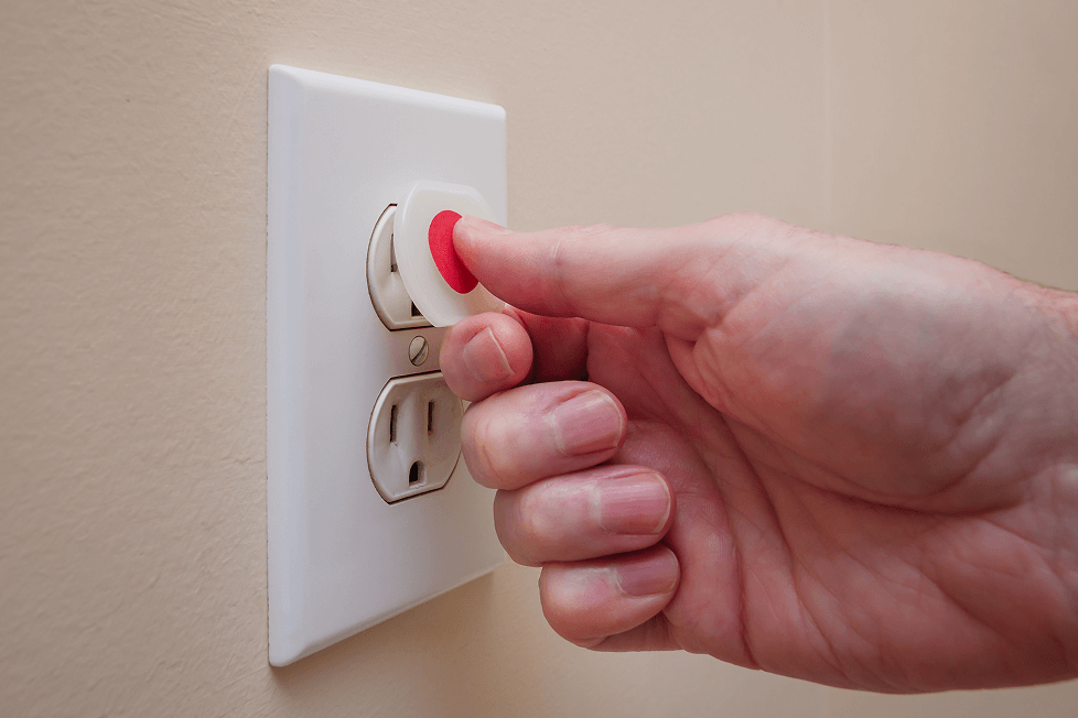 Gros plan d’une main insérant un bouchon de sécurité pour prise de courant à centre rouge dans une prise électrique, sur un mur beige.