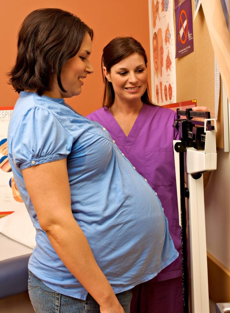A pregnant person in a blue shirt standing on a weighing scale, assisted by a healthcare professional in purple scrubs. Medical posters and charts are visible on the wall in the background.