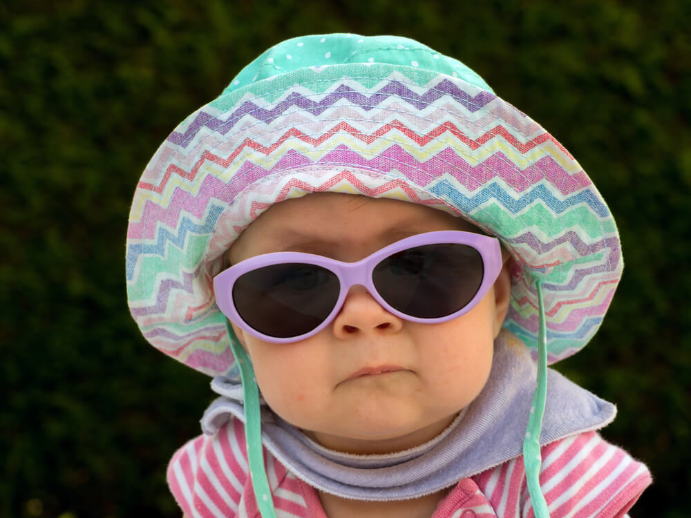 Enfant portant un chapeau coloré en zigzag dans des tons pastel de vert, mauve, rose et blanc. On voit des produits verts en arrière-plan.