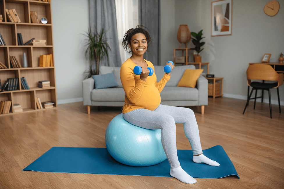 A pregnant person in a living room sits on an exercise ball while holding small dumbbells.