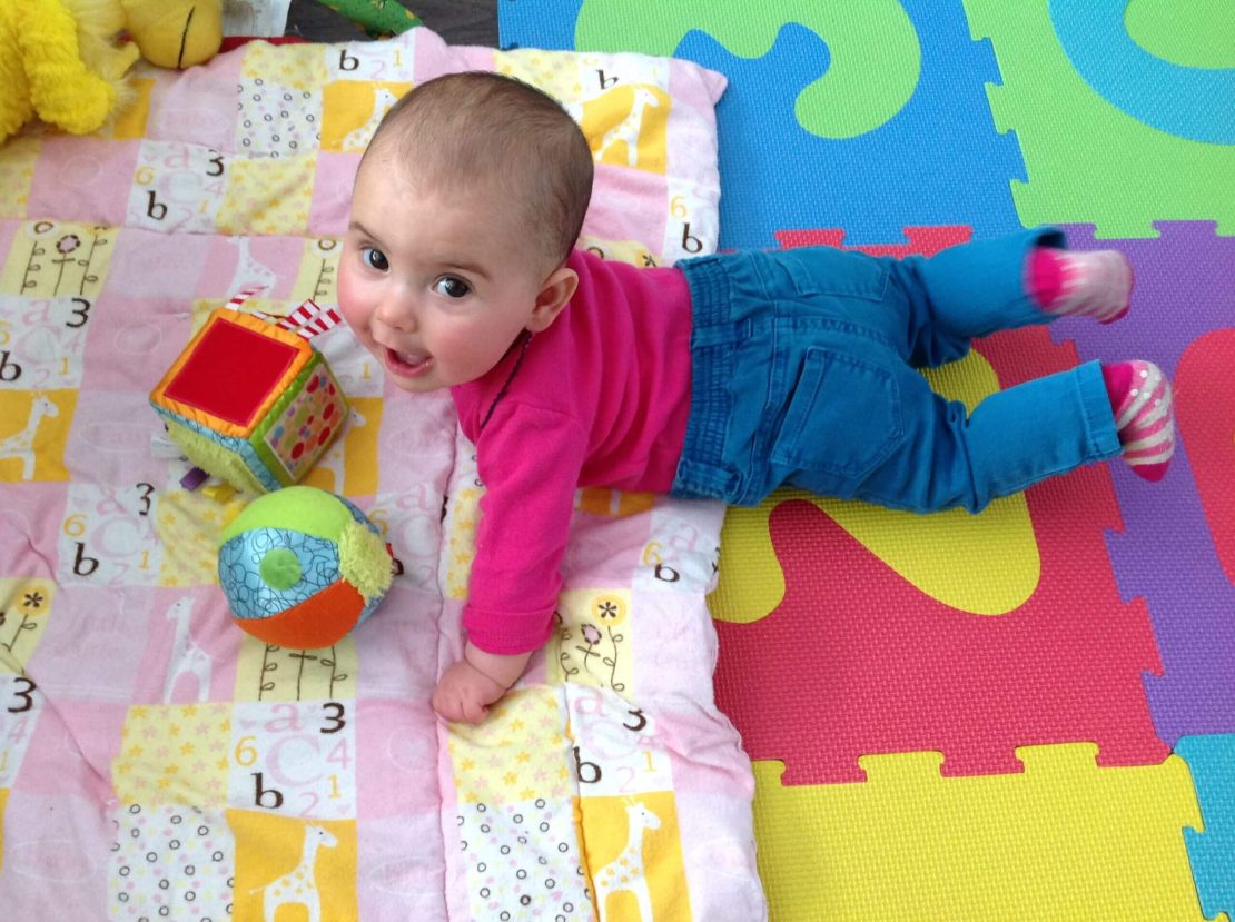 Bébé couché sur un tapis de jeu coloré, portant une chemise rose à manches longues, un pantalon bleu et des chaussettes rayées. À proximité se trouvent une boule multicolore et un jouet en forme de cube texturé. Le tapis présente des motifs comprenant des girafes, des chiffres et des lettres (a, b, c).