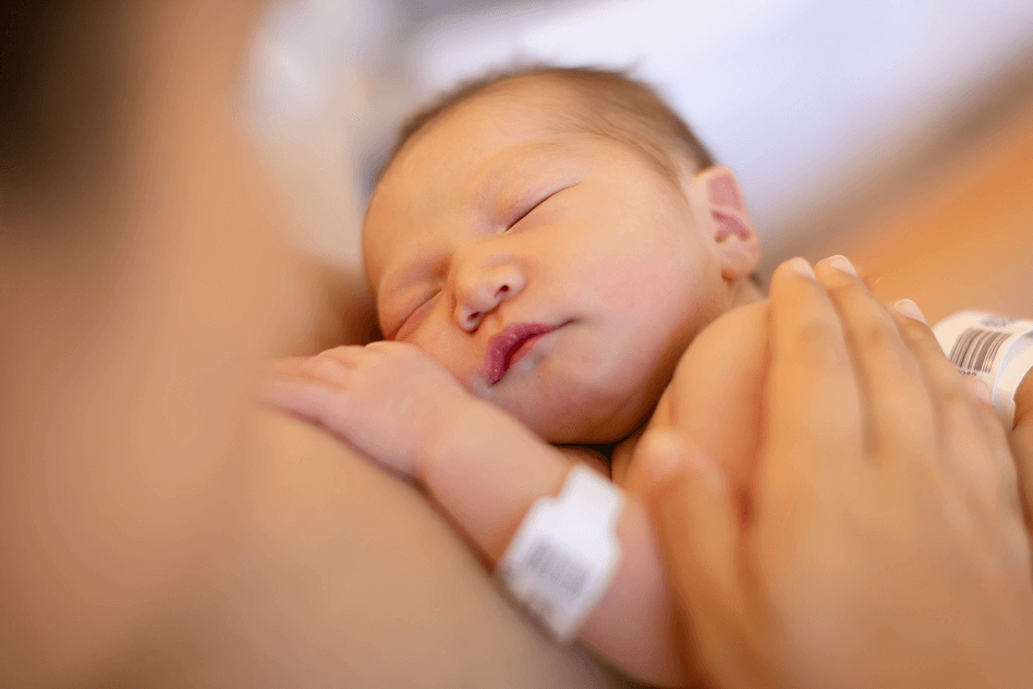A newborn baby with a hospital ID bracelet lies on an adult’s chest, with the adult’s hand resting gently on the baby’s back.