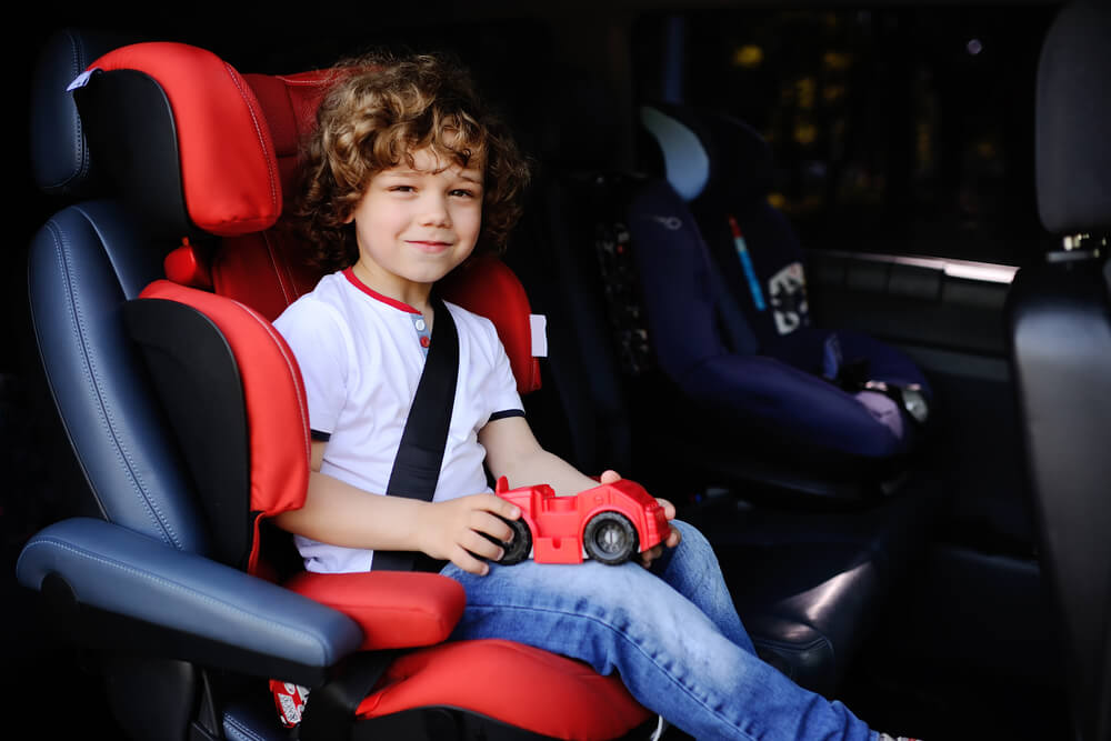 A child wearing a white shirt with red trim and blue jeans sits in a black and red car seat inside a vehicle, holding a red toy car. An empty car seat is visible in the background.