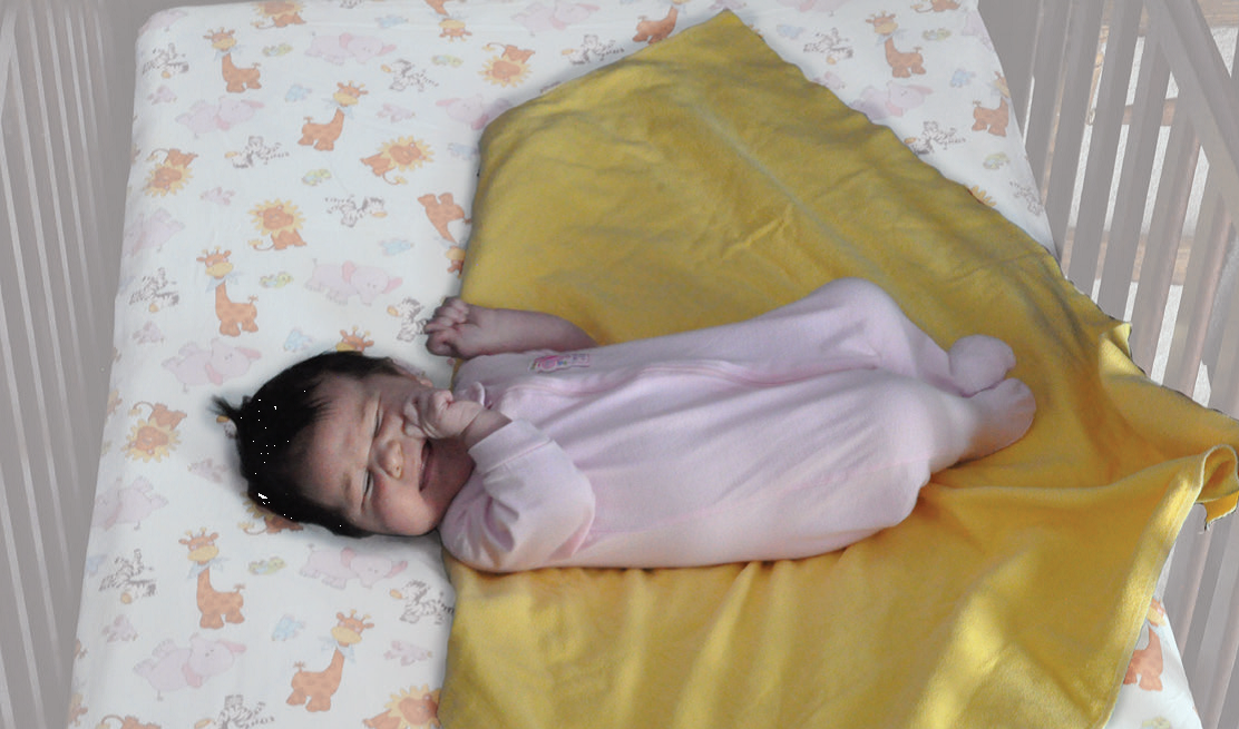 Un bébé aux cheveux foncés dans une grenouillère rose pâle sur une couverture jaune dont un coin est replié