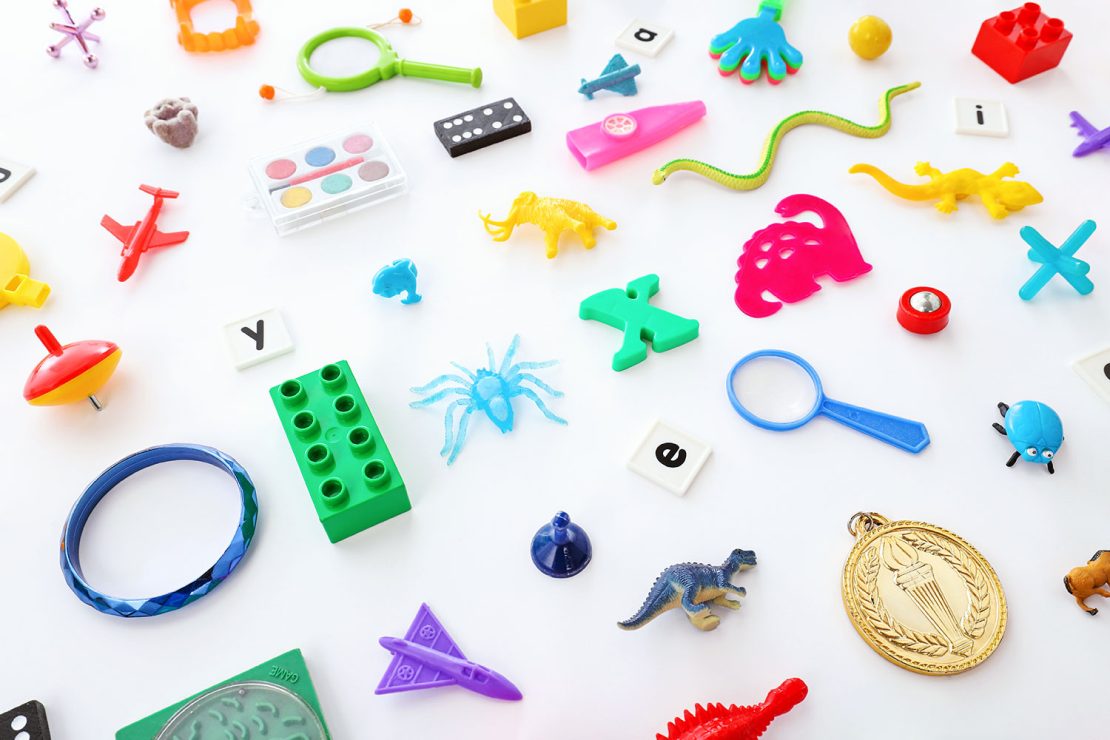Assortiment de petits jouets et objets colorés, y compris des animaux en plastique, des lettres, une loupe, un avion-jouet, une toupie et des blocs de construction, dispersés sur un fond blanc.