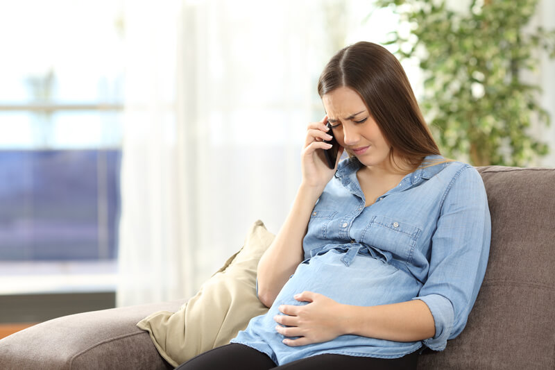 Personne assise sur un canapé, tenant un téléphone à l’oreille d’une main et reposant l’autre main sur son ventre de femme enceinte. Elle porte une chemise à boutons bleu pâle et un pantalon noir. Un oreiller beige se trouve à côté d’elle, et on voit des plantes vertes et une lumière naturelle vive en arrière-plan.