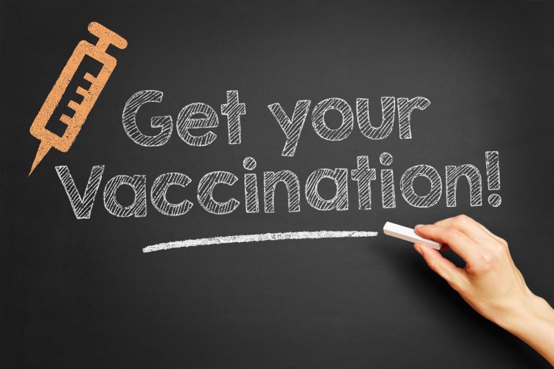 A blackboard with the text 'Get your Vaccination!' written in chalk. An illustration of a syringe is in the top left corner, and a hand holding a piece of chalk is visible on the right