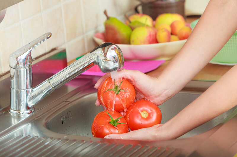 A person washing three tomatoes under a running faucet in a kitchen sink. In the background, a bowl filled with various fruits, including apples and pears, is visible.